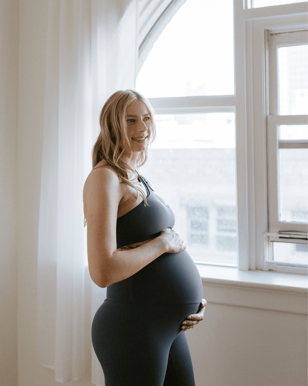 Pregnant woman wearing premium slate maternity leggings with a flattering scrunch butt and matching one-shoulder, nursing sports bra, smiling while standing near a bright window. Premium maternity athletic and activewear by AXK.