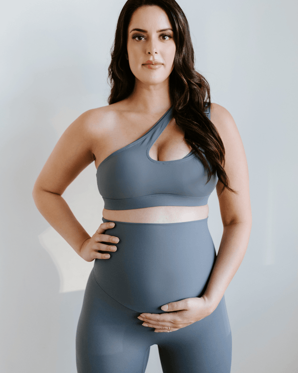 Pregnant woman wearing a blue one-shoulder, maternity, nursing sports bra and matching high-waisted maternity leggings, standing against a white background. Premium maternity activewear by AXK.