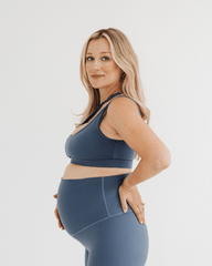 Side view of a pregnant woman wearing premium maternity leggings with a supportive high waist and a matching one-shoulder maternity and nursing sports bra in blue, standing against a white background. Premium maternity athletic and activewear by AXK.