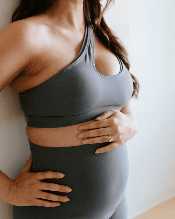 Close-up of a pregnant woman wearing a blue one-shoulder, maternity and nursing sports bra and leggings, with one hand resting on her baby bump. Premium maternity activewear by AXK.