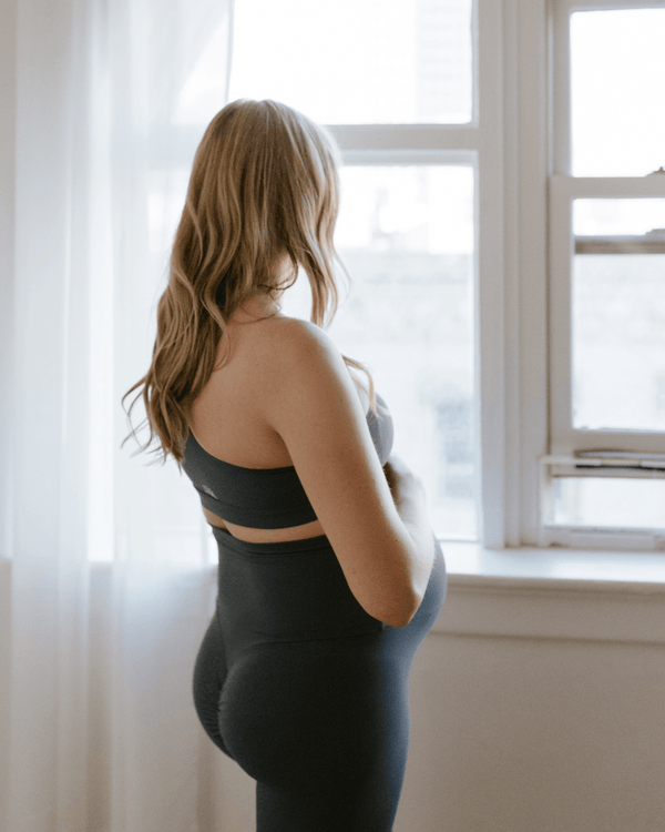 Pregnant woman wearing premium blue maternity leggings with a flattering scrunch butt and matching one-shoulder, nursing sports bra, standing in profile near a bright window. Premium maternity athletic and activewear by AXK.