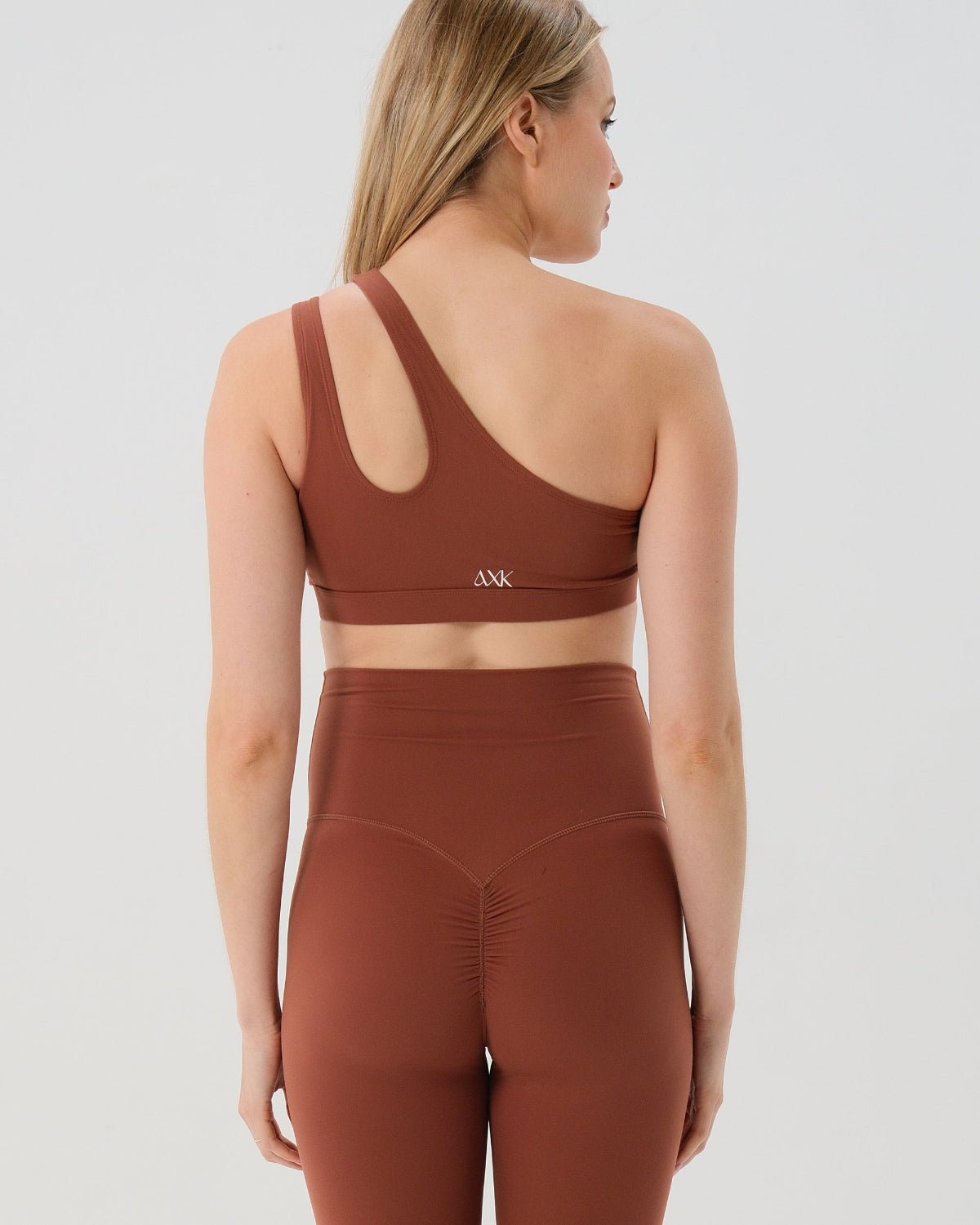 Pregnant woman wearing a brown one-shoulder maternity and nursing sports bra with matching high-waist leggings, standing against a white background. Premium maternity athletic and activewear by AXK - high quality petite, tall, and plus size maternity. 