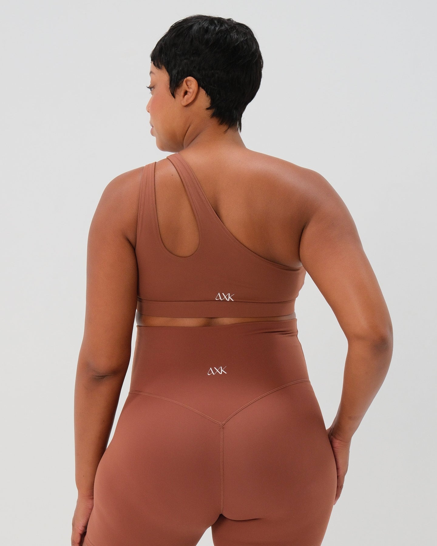 Pregnant woman in a one-shoulder brown maternity and nursing sports bra, wearing matching blue maternity leggings. Best selling 2026 premium maternity athletic and activewear by AXK, offered in regular and plus sizes, with glowing 5 star reviews. 