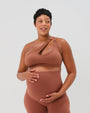 Pregnant woman wearing a brown one-shoulder maternity and nursing sports bra with matching high-waist leggings, standing against a white background. Premium maternity athletic and activewear by AXK - high quality petite, tall, and plus size maternity. 