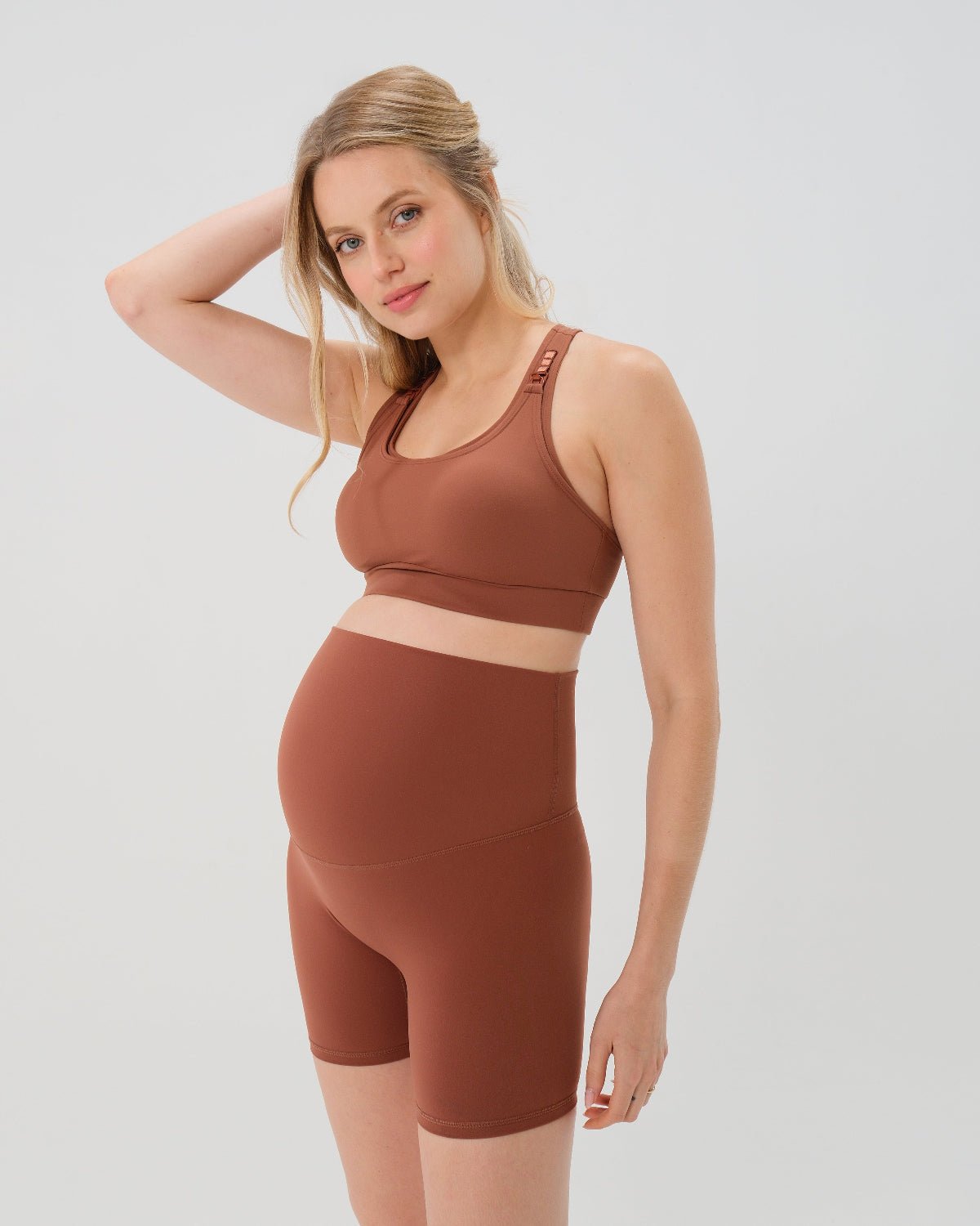 Pregnant woman wearing an AXK brown high-impact, racerback, maternity and nursing sports bra and matching high-waist maternity leggings. She has her hands cradling her belly standing in front of a white background. Premium maternity athletic and activewear by AXK - high quality petite, tall, and plus size maternity. 