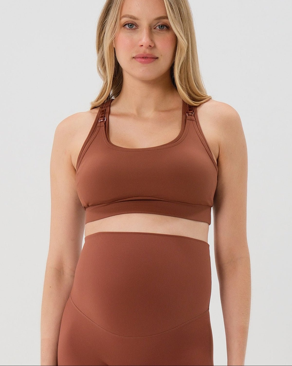 Pregnant woman wearing an AXK brown high-impact, racerback, maternity and nursing sports bra and matching high-waist maternity leggings. She has her hands cradling her belly standing in front of a white background. Premium maternity athletic and activewear by AXK - high quality petite, tall, and plus size maternity. 