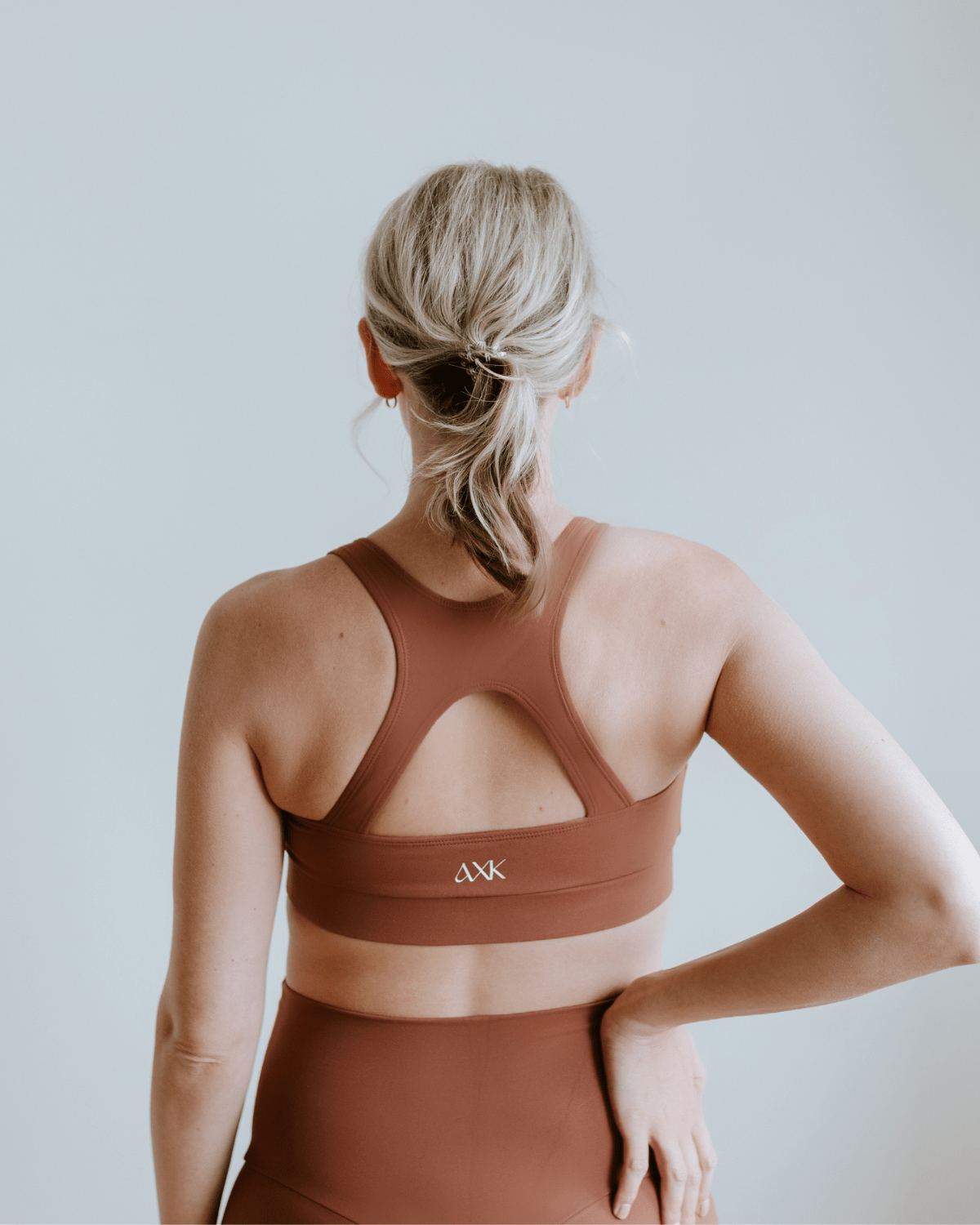 Back view of a pregnant woman wearing a brown high-impact, racerback, maternity and nursing sports bra and matching high-waist maternity leggings standing in front of a white background. Premium maternity athletic and activewear by AXK.