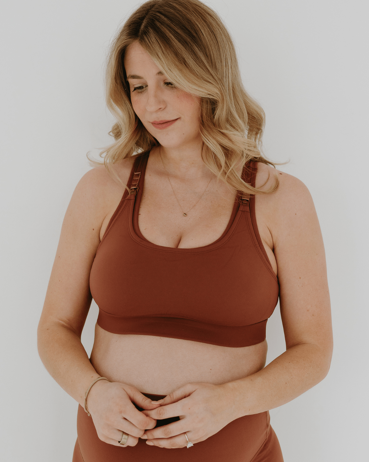 Close up view of a pregnant woman wearing a brown high-impact, racerback, maternity and nursing sports bra and matching high-waist maternity leggings. She has her hands cradling her belly standing in front of a white background. Premium maternity athletic and activewear by AXK.