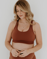 Pregnant woman wearing an AXK brown high-impact, racerback, maternity and nursing sports bra and matching high-waist maternity leggings. She has her hands cradling her belly standing in front of a white background. Premium maternity athletic and activewear by AXK - high quality petite, tall, and plus size maternity. 