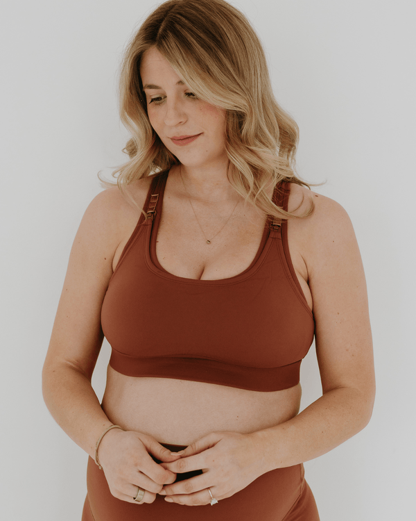Close up view of a pregnant woman wearing a brown high-impact, racerback, maternity and nursing sports bra and matching high-waist maternity leggings. She has her hands cradling her belly standing in front of a white background. Premium maternity athletic and activewear by AXK.