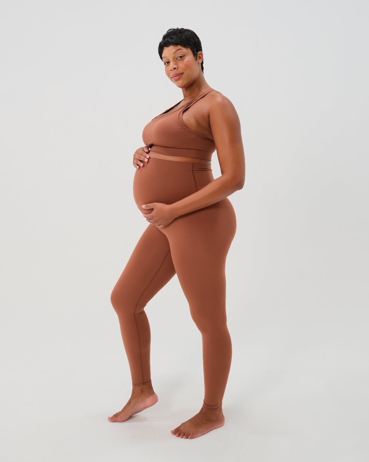 Pregnant woman wearing premium brown maternity leggings with a flattering scrunch butt and matching high-impact, racerback nursing sports bra, standing against a white background. Premium maternity athletic and activewear by AXK.