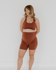 Pregnant woman wearing premium brown maternity bike shorts with a supportive high waist and matching high-impact, racerback maternity and nursing sports bra, standing against a white background. Premium maternity athletic and activewear by AXK.