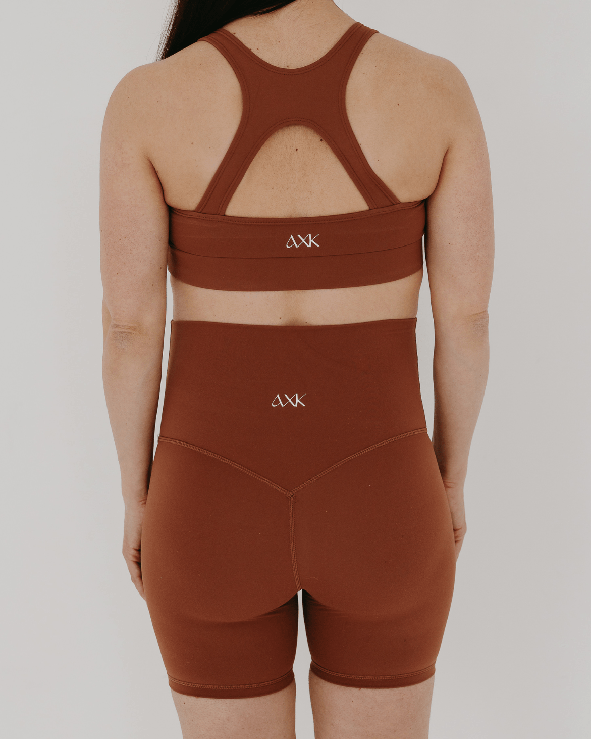 Back view of woman wearing premium brown AXK maternity bike shorts with a supportive high waist and matching high-impact, racerback maternity and nursing sports bra, both featuring the AXK logo, standing against a white background. Premium maternity athletic and activewear by AXK - high quality petite, tall, and plus size maternity. 