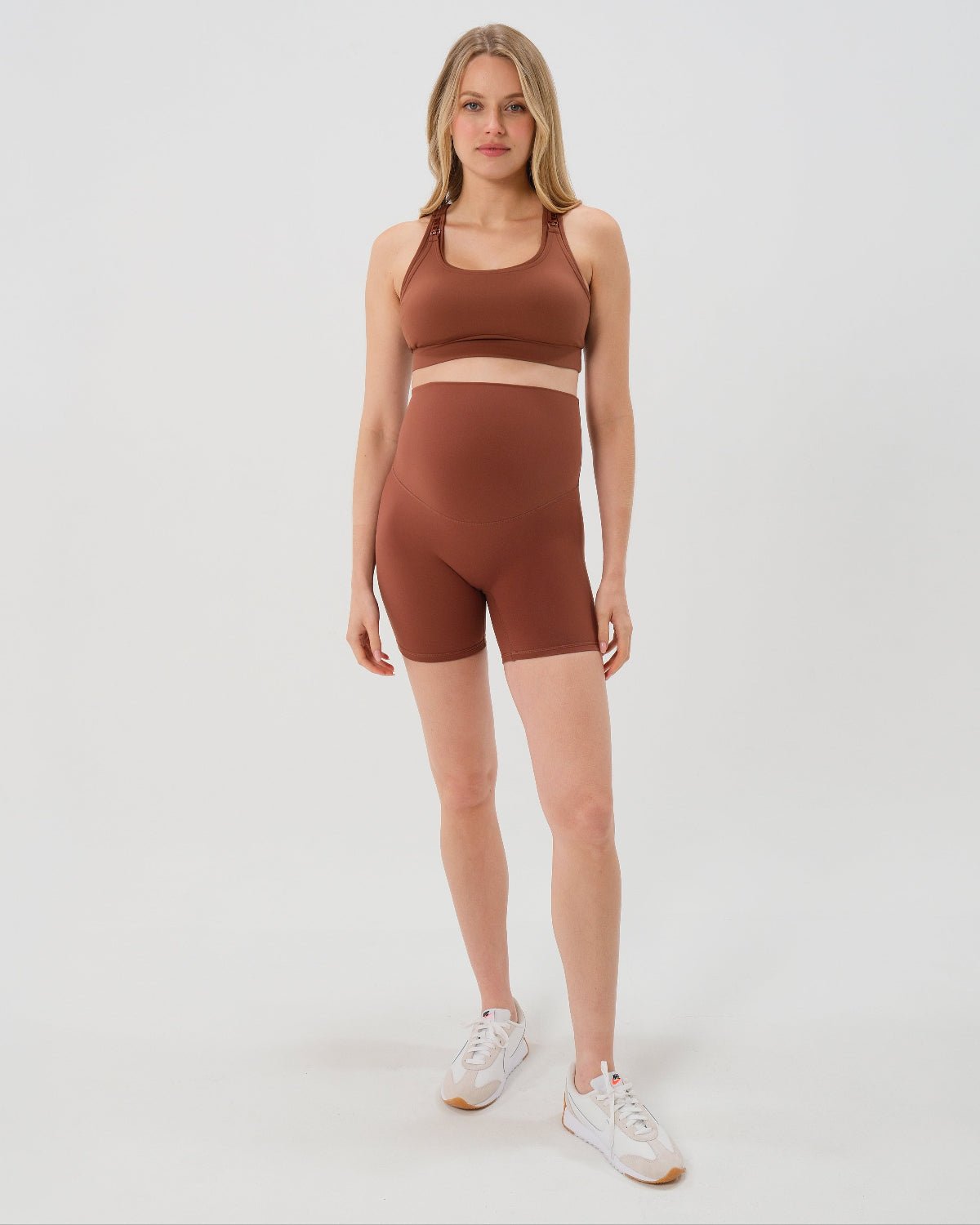 Pregnant woman wearing premium brown maternity bike shorts with a supportive high waist and matching high-impact, racerback maternity and nursing sports bra, standing against a white background. Premium maternity athletic and activewear by AXK - high quality petite, tall, and plus size maternity. 
