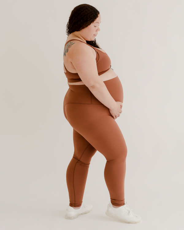 Side view of a pregnant woman wearing premium brown maternity leggings with a supportive high waist and matching high-impact racerback maternity and nursing sports bra, standing against a white background. Premium maternity athletic and activewear by AXK.