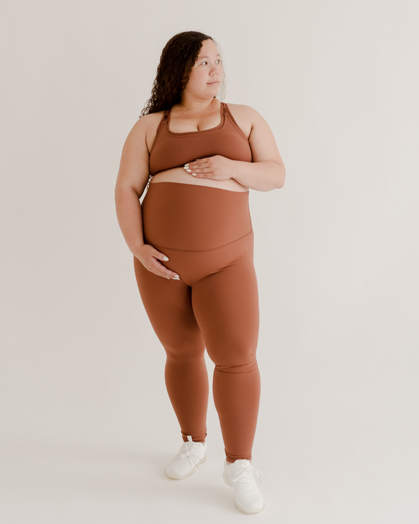 Pregnant woman wearing premium brown maternity leggings with a supportive high waist and matching high-impact racerback maternity and nursing sports bra, standing against a white background. Premium maternity athletic and activewear by AXK.