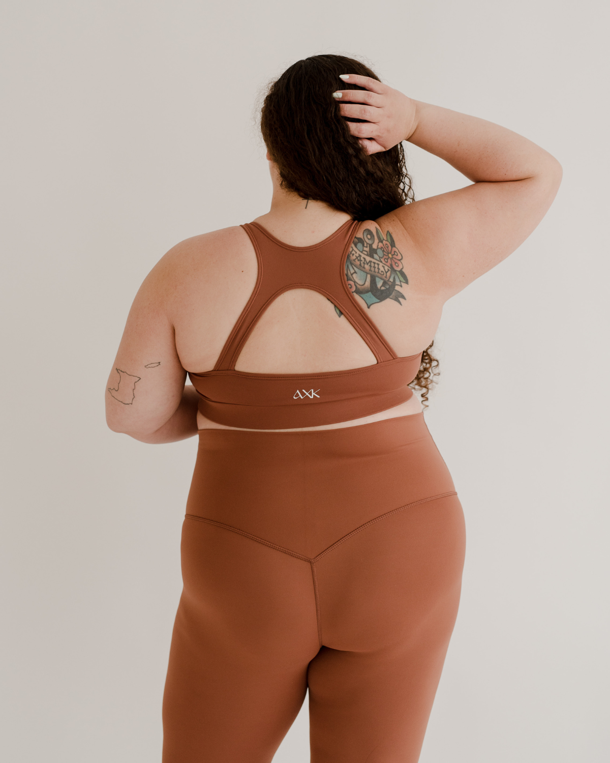 Close up back view of a pregnant woman wearing premium brown maternity leggings with a supportive high waist and matching high-impact racerback maternity and nursing sports bra, standing against a white background. Premium maternity athletic and activewear by AXK.