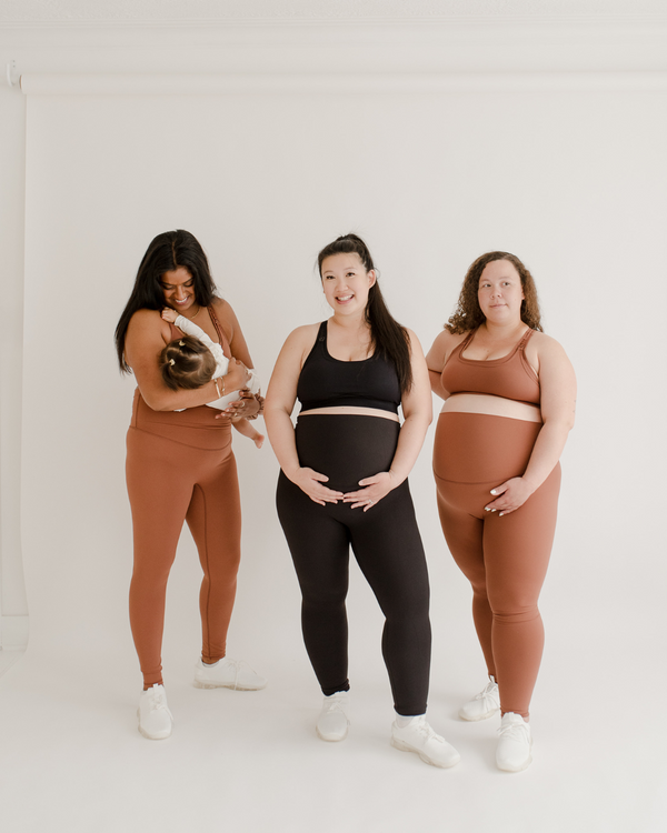 Three women wearing premium maternity leggings with supportive high waists and matching high-impact racerback maternity and nursing sports bras in brown and black, standing on a white background, with one mother holding her baby. Premium maternity athletic and activewear by AXK.