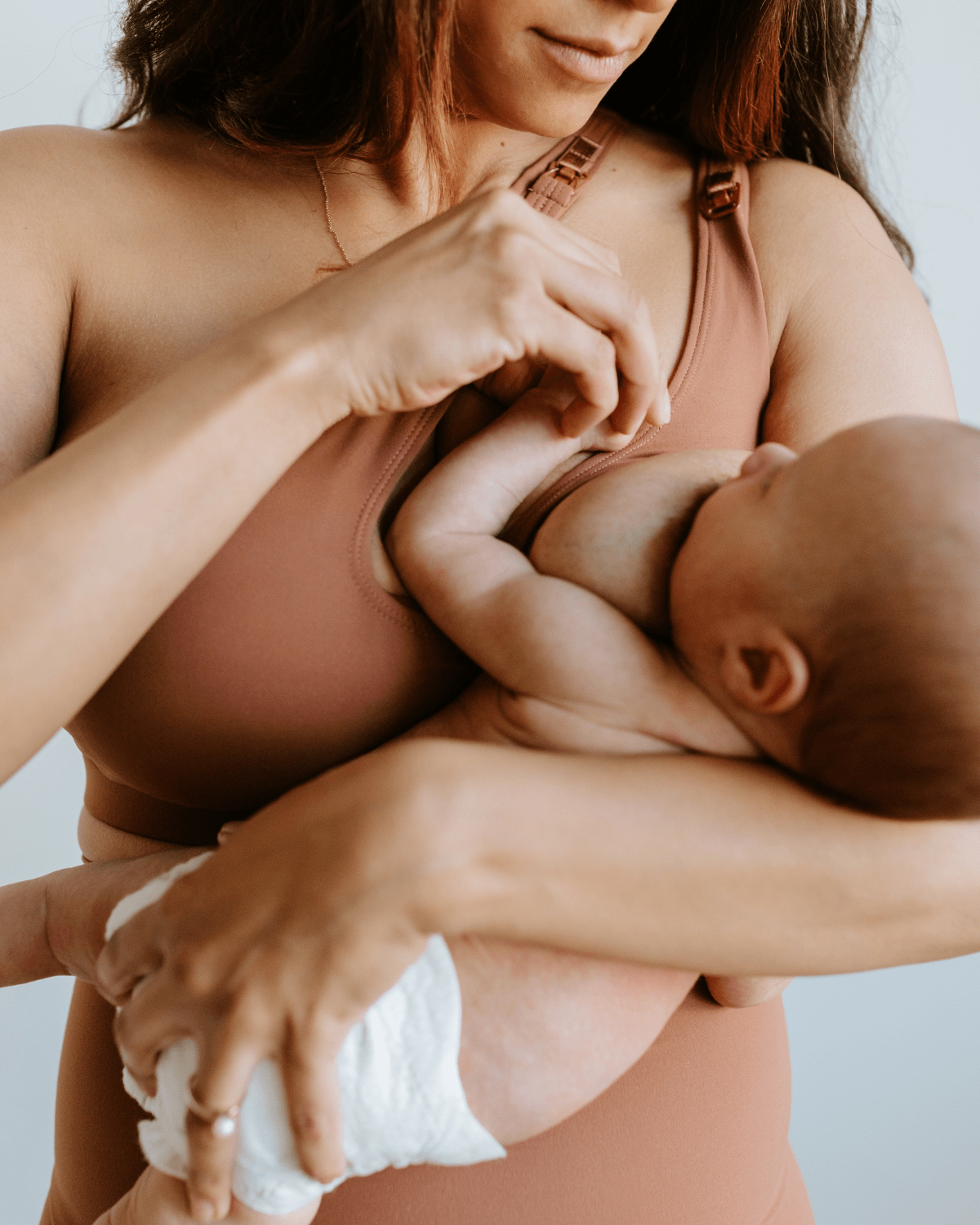 Woman nursing a baby while wearing a brown one-shoulder maternity and nursing sports bra. Premium maternity activewear by AXK.