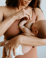 Woman nursing a baby while wearing a brown one-shoulder maternity and nursing sports bra. Premium maternity activewear by AXK.