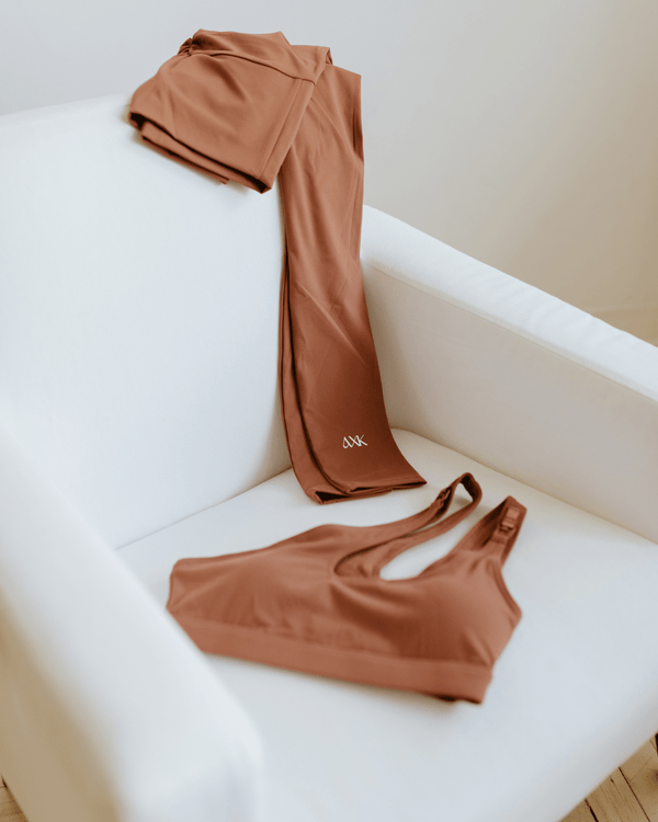 Brown one-shoulder, maternity sports bra and maternity leggings displayed on a white armchair. AXK premium maternity activewear.