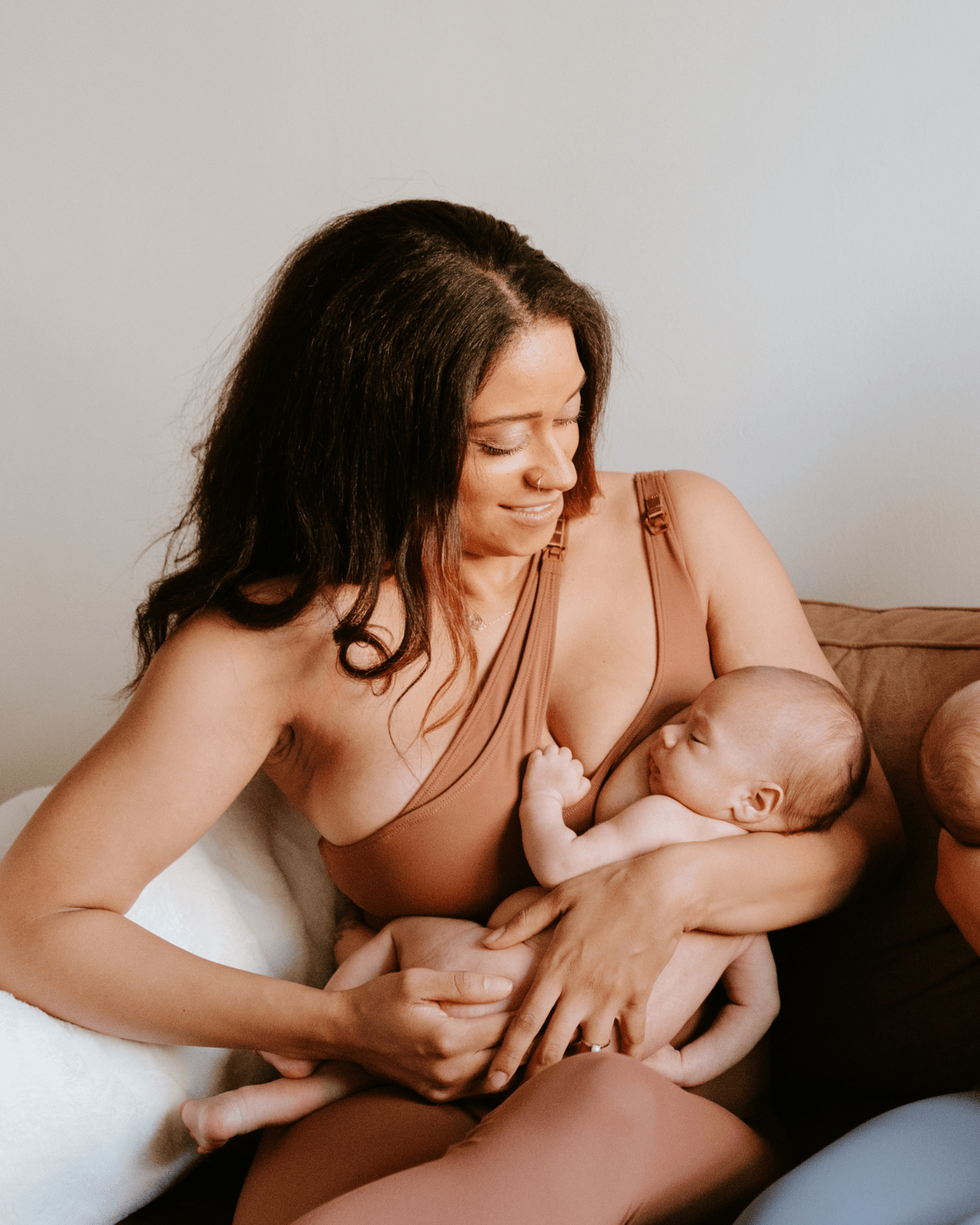 Mother wearing a brown one-shoulder, maternity and nursing sports bra and matching leggings, while holding and nursing a newborn baby. AXK premium maternity activewear