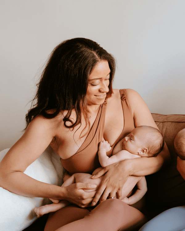 Mother wearing a brown one-shoulder, maternity and nursing sports bra and matching leggings, while holding and nursing a newborn baby. AXK premium maternity activewear