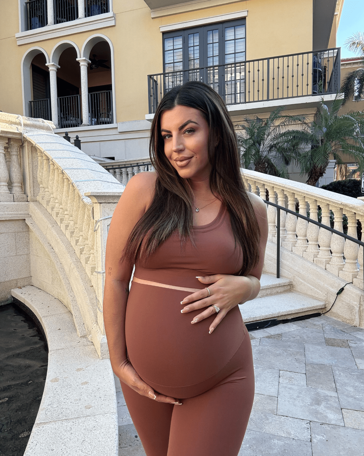 Plus size pregnant woman wearing premium brown maternity leggings with a flattering scrunch butt and matching high-impact, racerback nursing sports bra, standing against a white background. Premium maternity athletic and activewear by AXK - tall, petite, and plus size friendly.