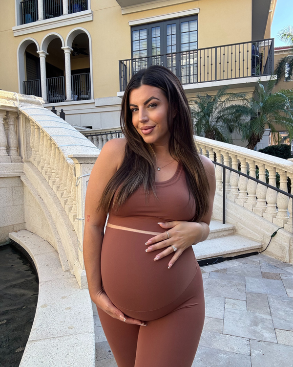 Pregnant woman wearing premium brown maternity leggings with a flattering scrunch butt and matching high-impact, racerback nursing sports bra, standing outdoors in an urban setting. Premium maternity athletic and activewear by AXK.