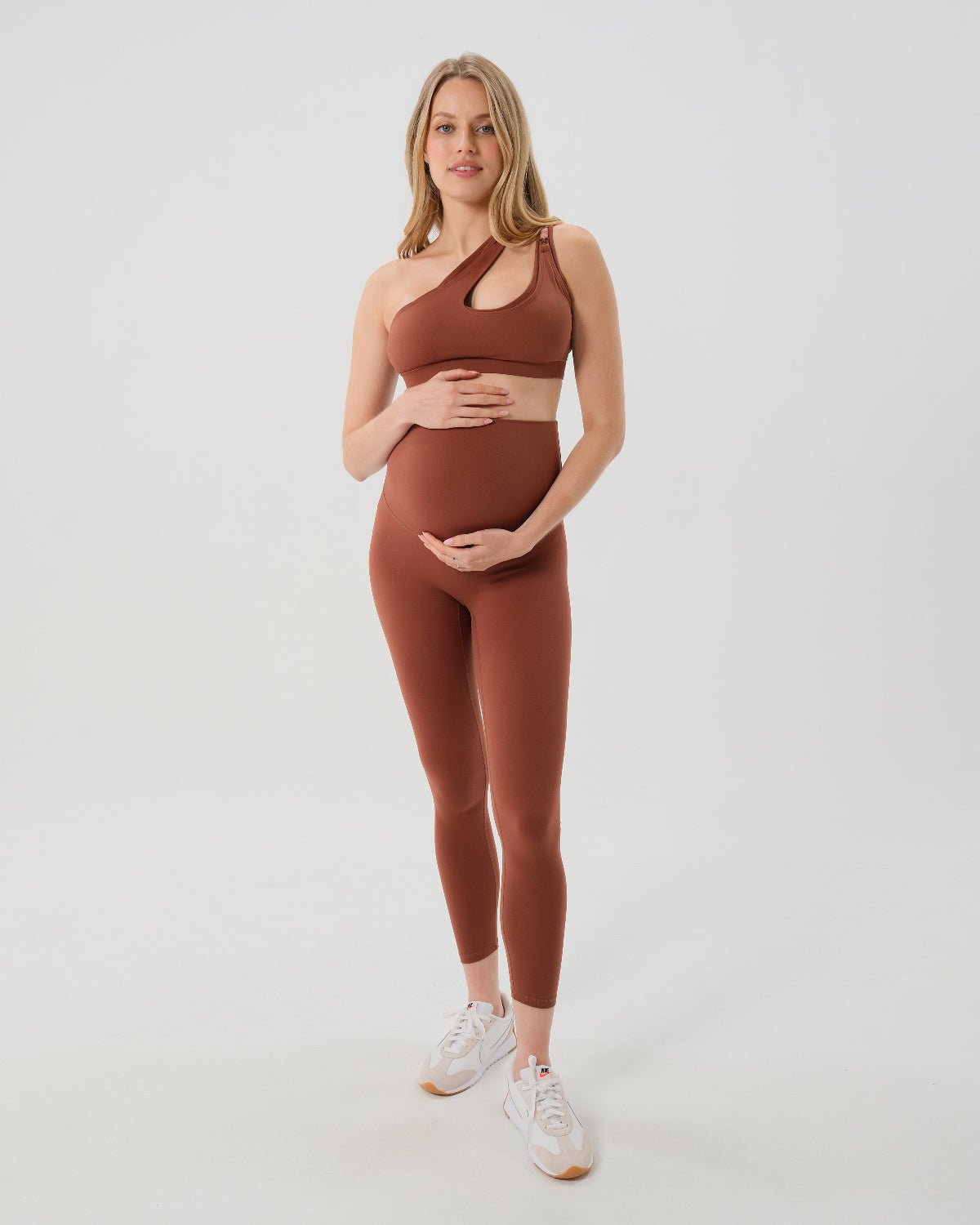 Pregnant woman wearing premium brown maternity leggings with a flattering scrunch butt and matching high-impact, racerback nursing sports bra, standing against a white background. Premium maternity athletic and activewear by AXK.