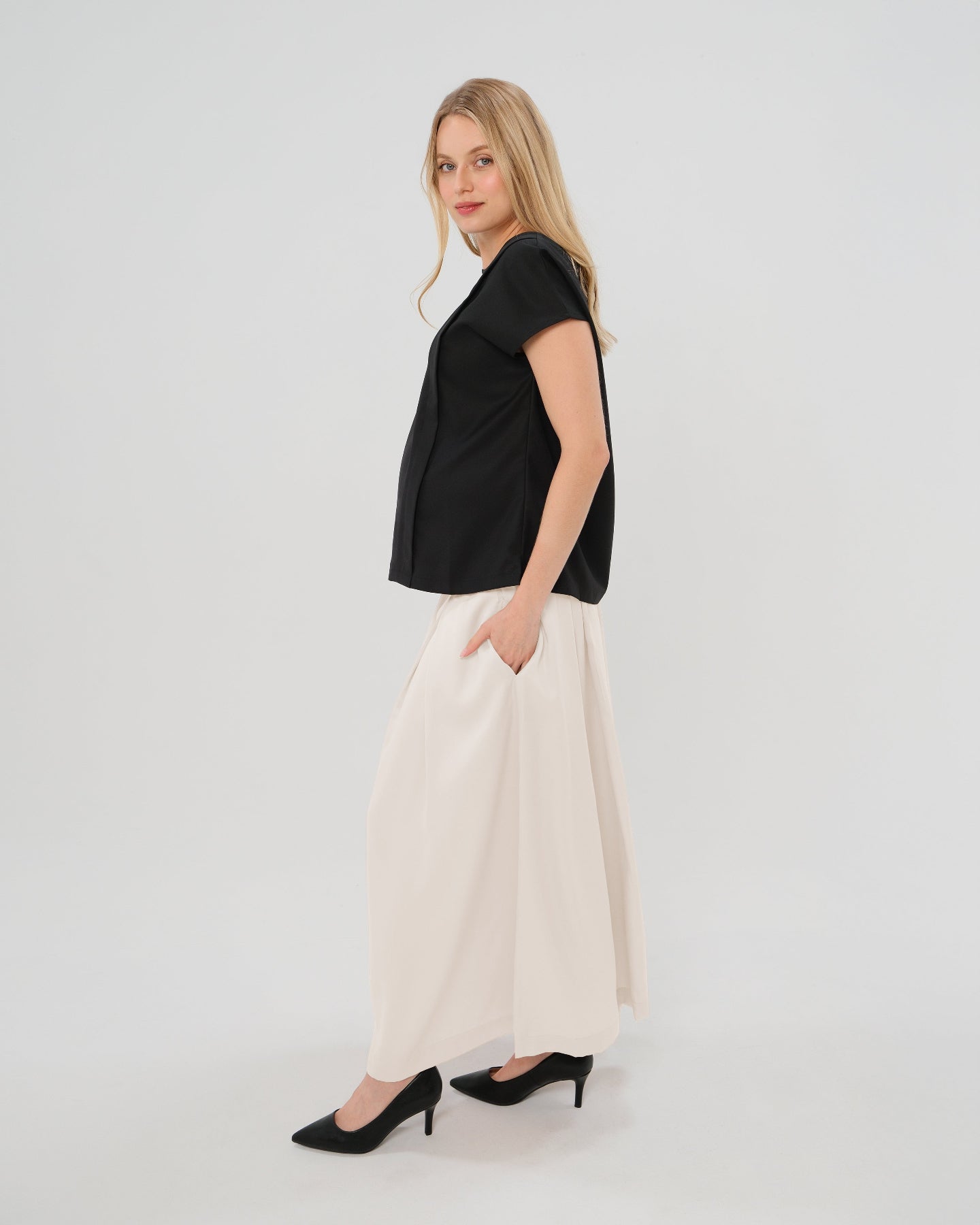 Pregnant woman wearing a modest off-white cream pleated designer maternity maxi maternity skirt in luxurious TENCEL satin by MARION. This luxury skirt can be worn over or under your belly bump. It fastens with a zip, and features deep functional pockets and a flexible elastic waistband for comfort. This refined, timeless style epitomizes quiet luxury. Perfect for maternity work wear, wedding guest dress, occasion wear, pregnancy street style, and baby shower outfits. Petite and tall friendly.