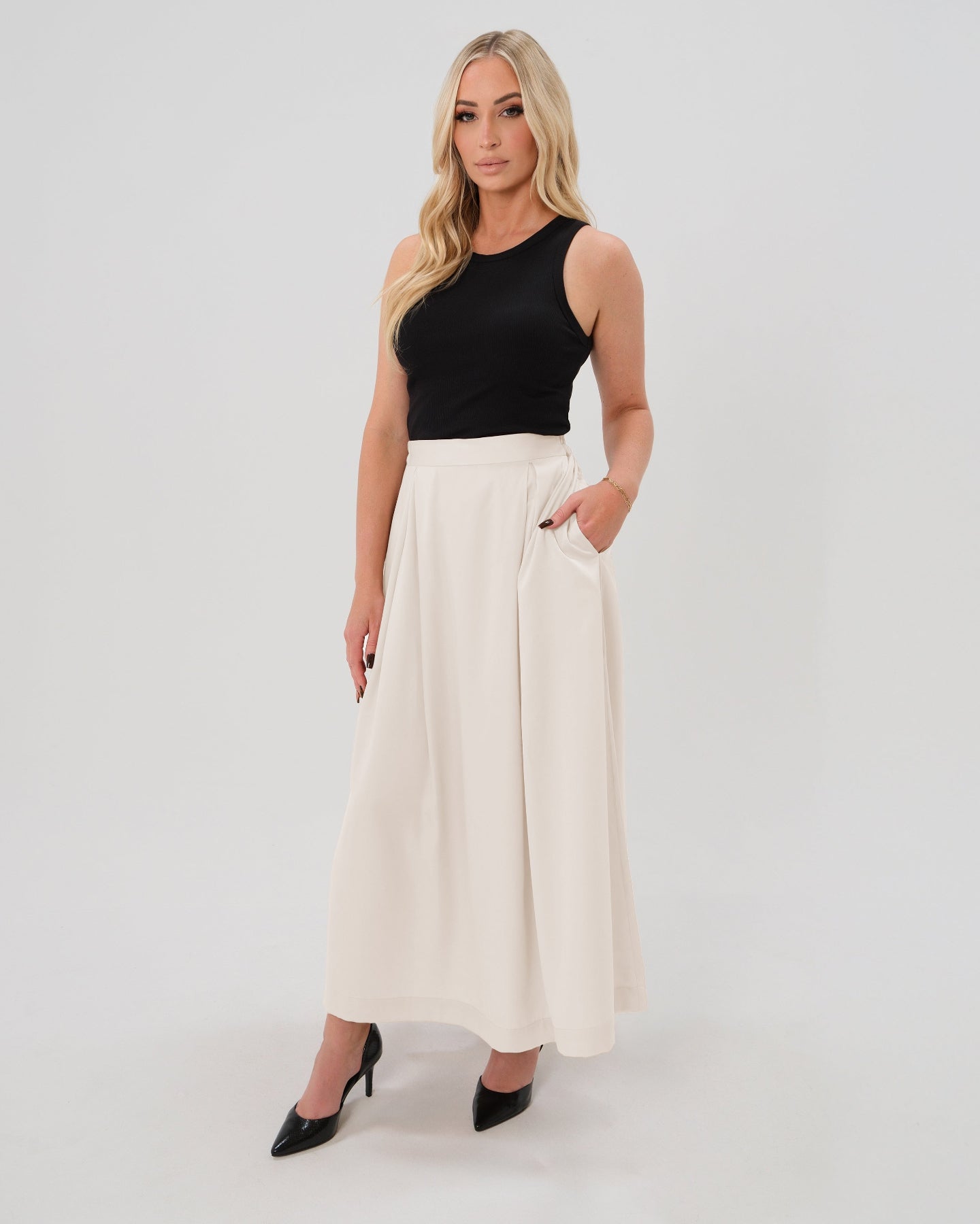 Pregnant woman wearing a modest off-white cream pleated designer maternity maxi maternity skirt in luxurious TENCEL satin by MARION. This luxury skirt can be worn over or under your belly bump. It fastens with a zip, and features deep functional pockets and a flexible elastic waistband for comfort. This refined, timeless style epitomizes quiet luxury. Perfect for maternity work wear, wedding guest dress, occasion wear, pregnancy street style, and baby shower outfits. Petite and tall friendly.
