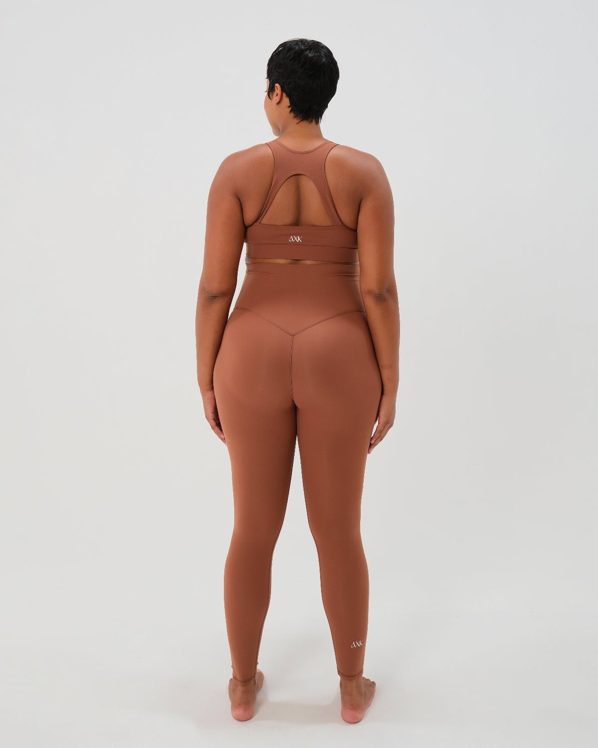 Pregnant woman wearing premium brown maternity leggings with a flattering scrunch butt and matching high-impact, racerback nursing sports bra, standing outdoors in an urban setting. Premium maternity athletic and activewear by AXK.