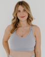 Smiling woman wearing a light-gray, high-impact, racerback maternity and nursing bra with clip-down straps, standing against a white background. AXK premium maternity activewear.