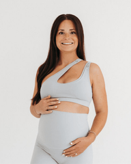 Pregnant woman wearing a premium light-gray one-shoulder, low-impact maternity and nursing sports bra with clip-down access, paired with high-waist maternity leggings, standing against a light background. Premium maternity athletic and activewear by AXK.