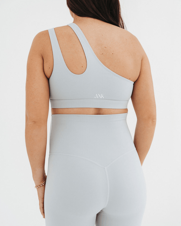 Back view of a pregnant woman wearing a premium light-gray one-shoulder, low-impact maternity and nursing sports bra with clip-down access, paired with high-waist maternity leggings, standing against a light background. Premium maternity athletic and activewear by AXK.