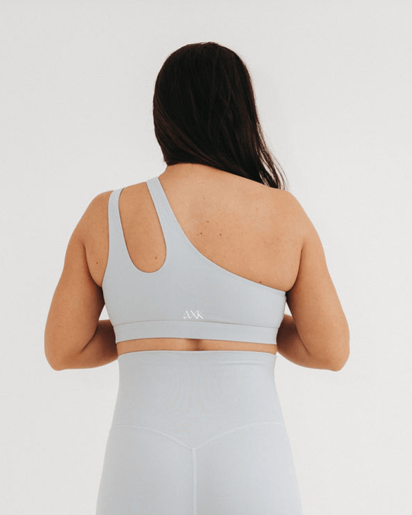 Back view of a pregnant woman wearing a premium light-gray one-shoulder, low-impact maternity and nursing sports bra with clip-down access, paired with high-waist maternity leggings, standing against a light background. Premium maternity athletic and activewear by AXK.