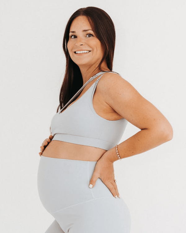 Side view of a pregnant woman wearing a premium light-gray one-shoulder, low-impact maternity and nursing sports bra with clip-down access, paired with high-waist maternity leggings, standing against a light background. Premium maternity athletic and activewear by AXK.