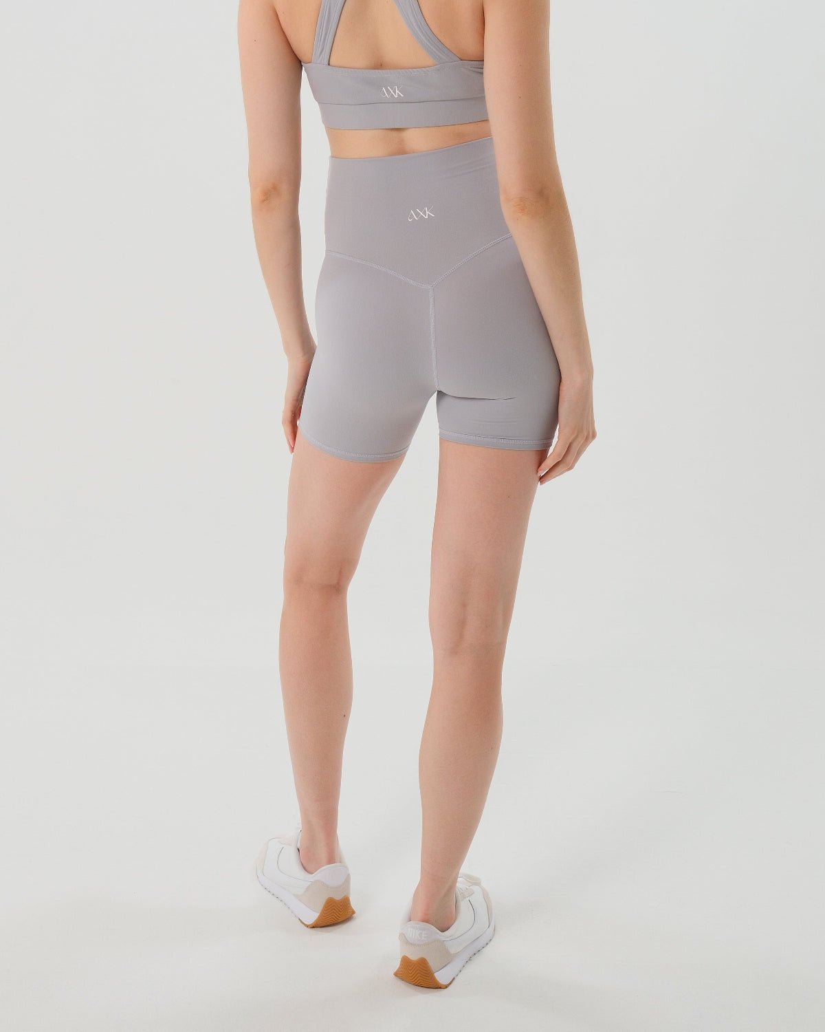 Pregnant woman wearing premium light grey maternity bike shorts with a supportive high waist and matching high-impact, racerback maternity and nursing sports bra, standing against a white background. Premium maternity athletic and activewear by AXK. Back view.