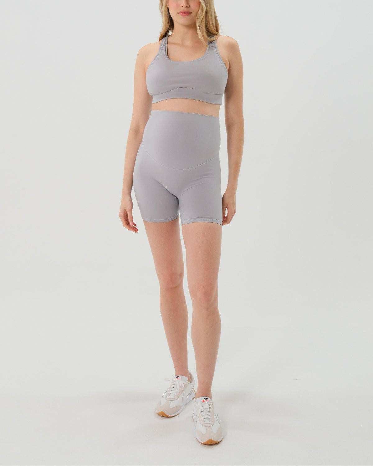 Pregnant woman wearing premium light grey maternity bike shorts with a supportive high waist and matching high-impact, racerback maternity and nursing sports bra, standing against a white background. Premium maternity athletic and activewear by AXK.