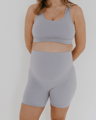 Pregnant woman wearing premium light grey maternity bike shorts with a supportive high waist and matching high-impact, racerback maternity and nursing sports bra, standing against a white background. Premium maternity athletic and activewear by AXK.