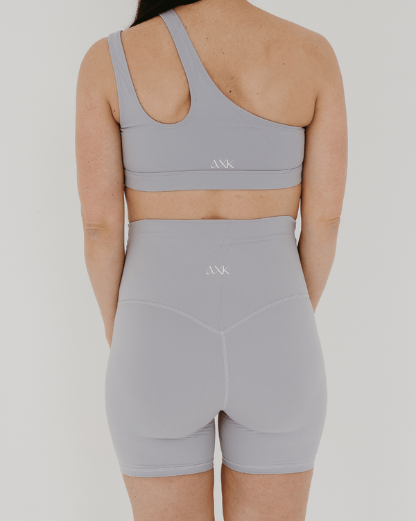 Back view of a pregnant woman wearing premium light grey maternity bike shorts with a supportive high waist and matching high-impact, racerback maternity and nursing sports bra, standing against a white background. Premium maternity athletic and activewear by AXK.