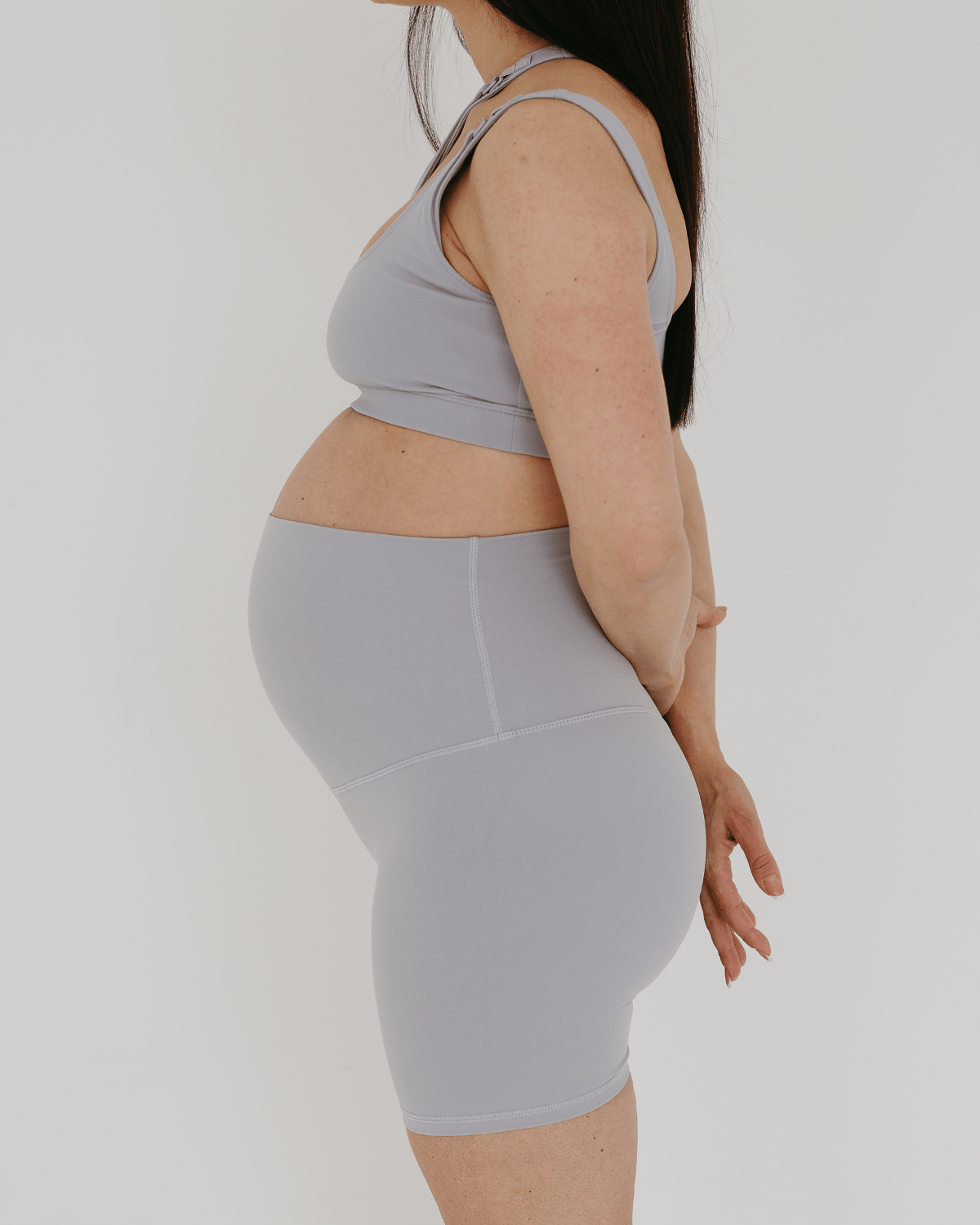 Side view of a pregnant woman wearing premium light grey maternity bike shorts with a supportive high waist and matching high-impact, racerback maternity and nursing sports bra, standing against a white background. Premium maternity athletic and activewear by AXK.