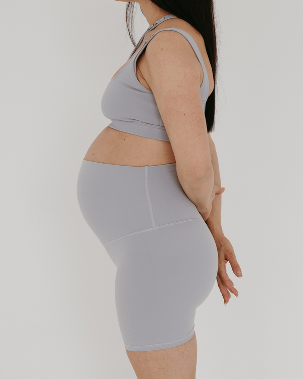 Side view of a pregnant woman wearing premium light grey maternity bike shorts with a supportive high waist and matching high-impact, racerback maternity and nursing sports bra, standing against a white background. Premium maternity athletic and activewear by AXK.