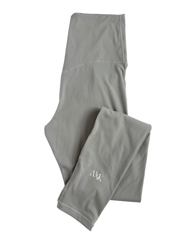 Flat lay of premium light grey maternity leggings with a flattering V-seam back and AXK logo detail on the ankle, displayed on a white background. Premium maternity athletic and activewear by AXK.