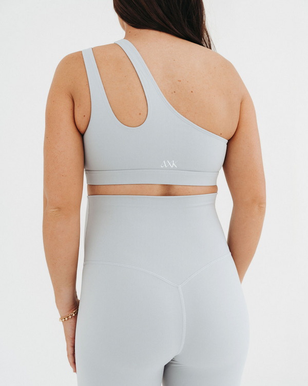 Close up back view of a pregnant woman wearing premium light grey maternity leggings with a flattering V-seam back and matching one-shoulder, low-impact, maternity and nursing sports bra, standing against a white background. Premium maternity athletic and activewear by AXK.