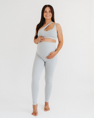 Pregnant woman wearing premium light grey maternity leggings with a flattering V-seam back and matching one-shoulder, low-impact, maternity and nursing sports bra, standing against a white background. Premium maternity athletic and activewear by AXK.