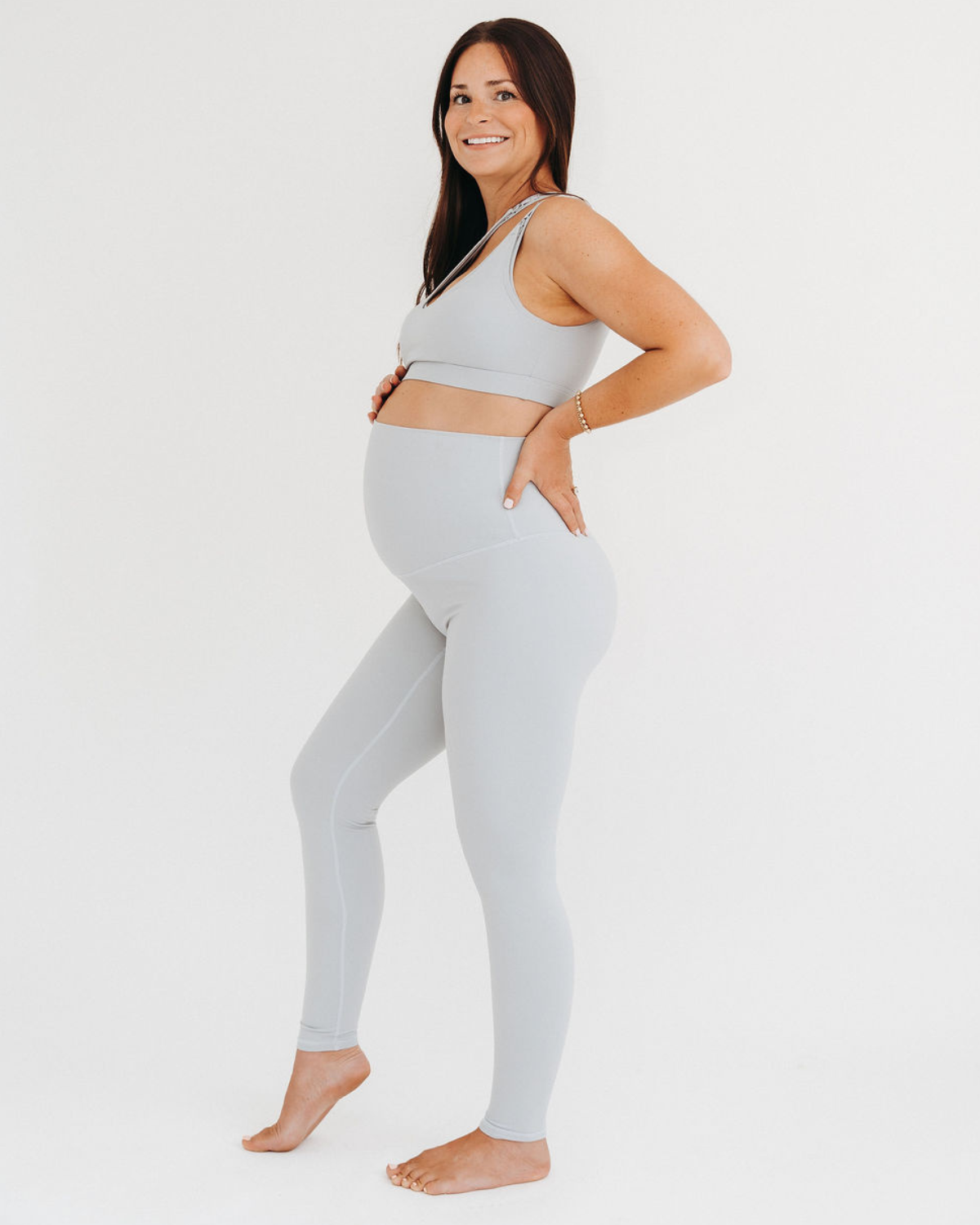 Side view of a pregnant woman wearing premium light grey maternity leggings with a flattering V-seam back and matching one-shoulder, low-impact, maternity and nursing sports bra, standing against a white background. Premium maternity athletic and activewear by AXK.