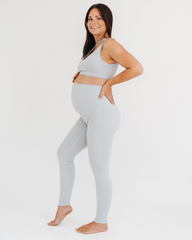 Side view of a pregnant woman wearing premium light grey maternity leggings with a flattering V-seam back and matching one-shoulder, low-impact, maternity and nursing sports bra, standing against a white background. Premium maternity athletic and activewear by AXK.