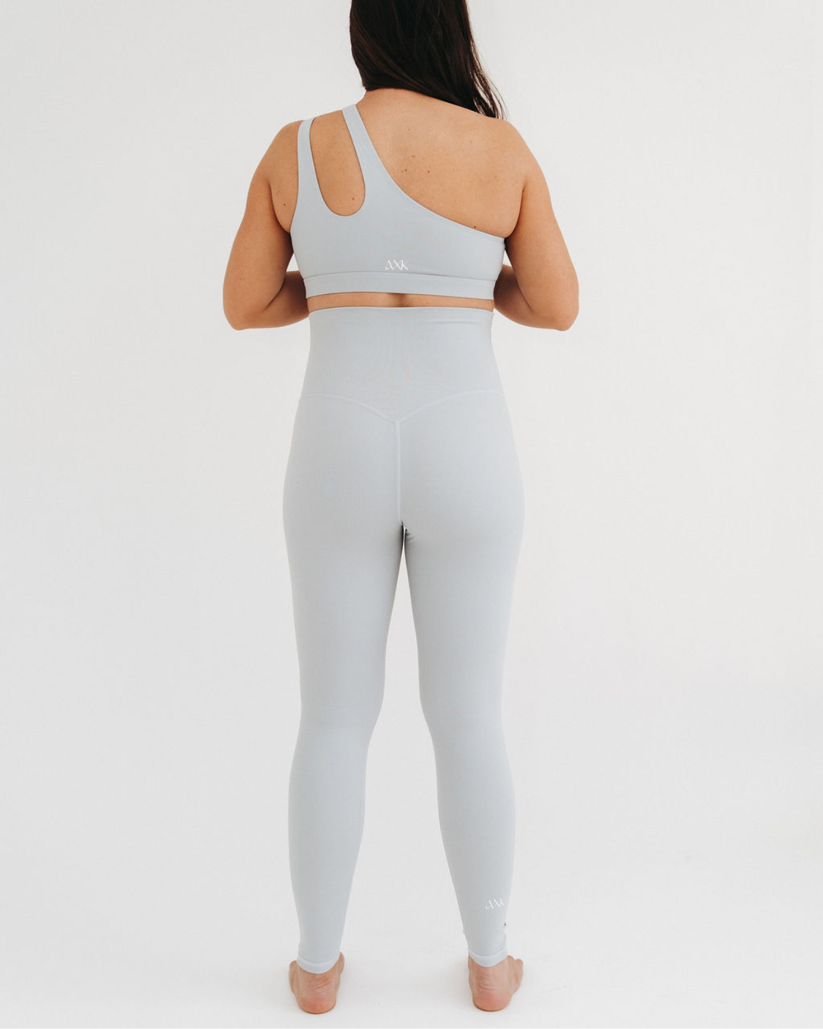 Back view of a pregnant woman wearing premium light grey maternity leggings with a flattering V-seam back and matching one-shoulder, low-impact, maternity and nursing sports bra, standing against a white background. Premium maternity athletic and activewear by AXK.